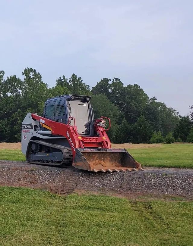 A red and grey tracked skid steer loader parked on a gravel path in a grassy field with trees in the background.