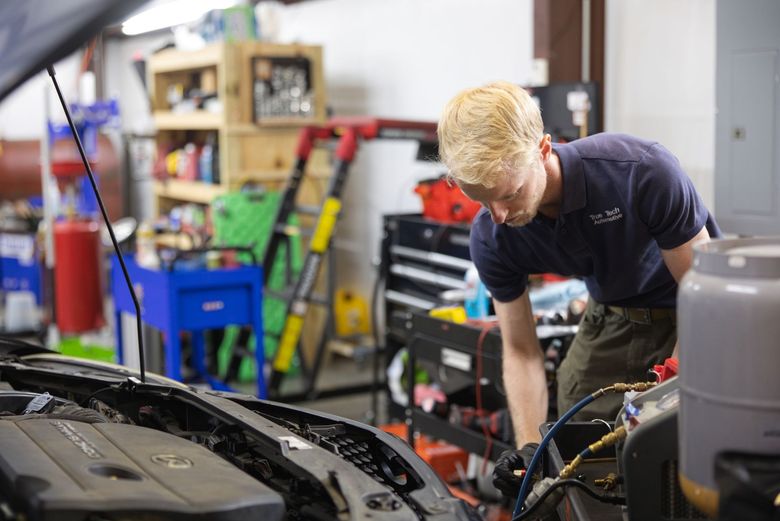 Mechanic working under an open car hood in a garage with tools and shelves in the background