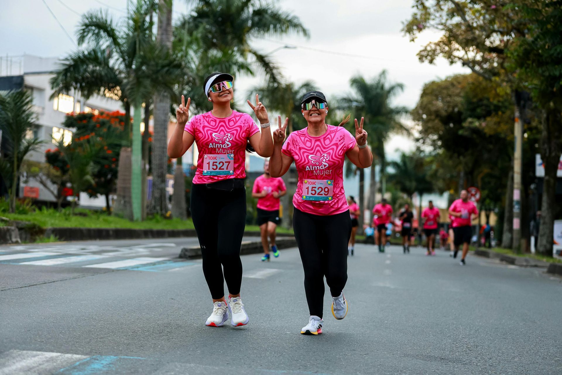 Two runners in pink shirts race along a palm-lined street, with more runners behind them.