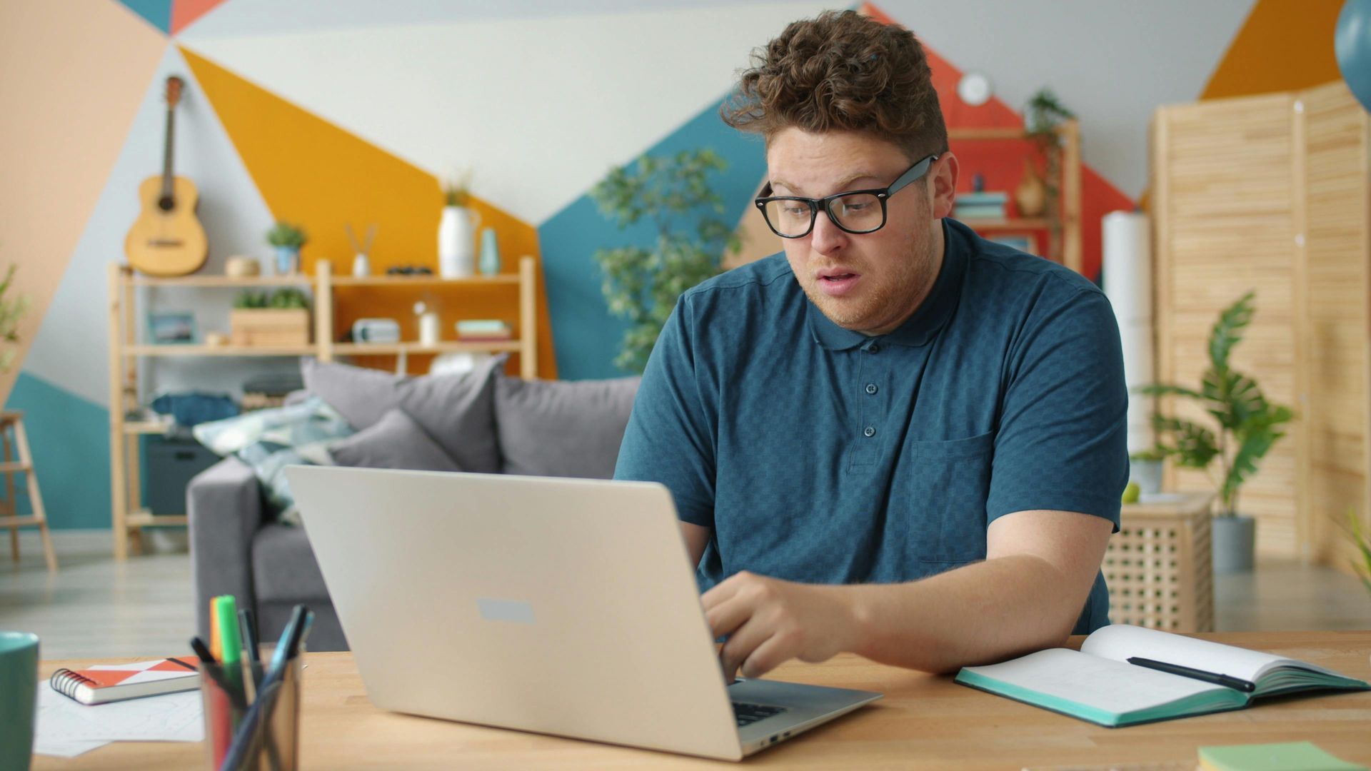 Person working on a laptop at a desk in a colorful home office, with a notebook nearby.