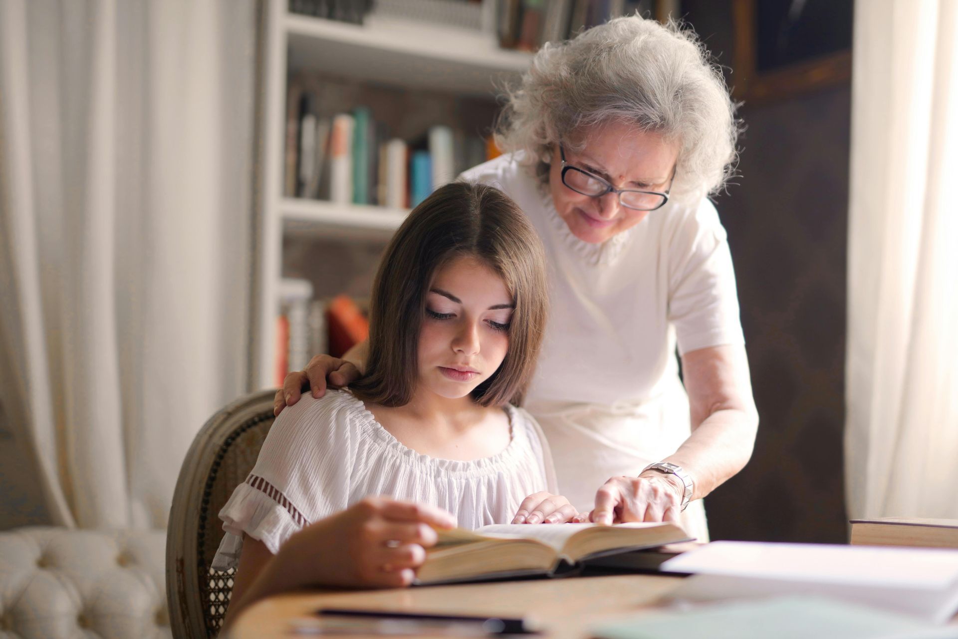 Older woman helps a girl read a book at a table in a cozy home library