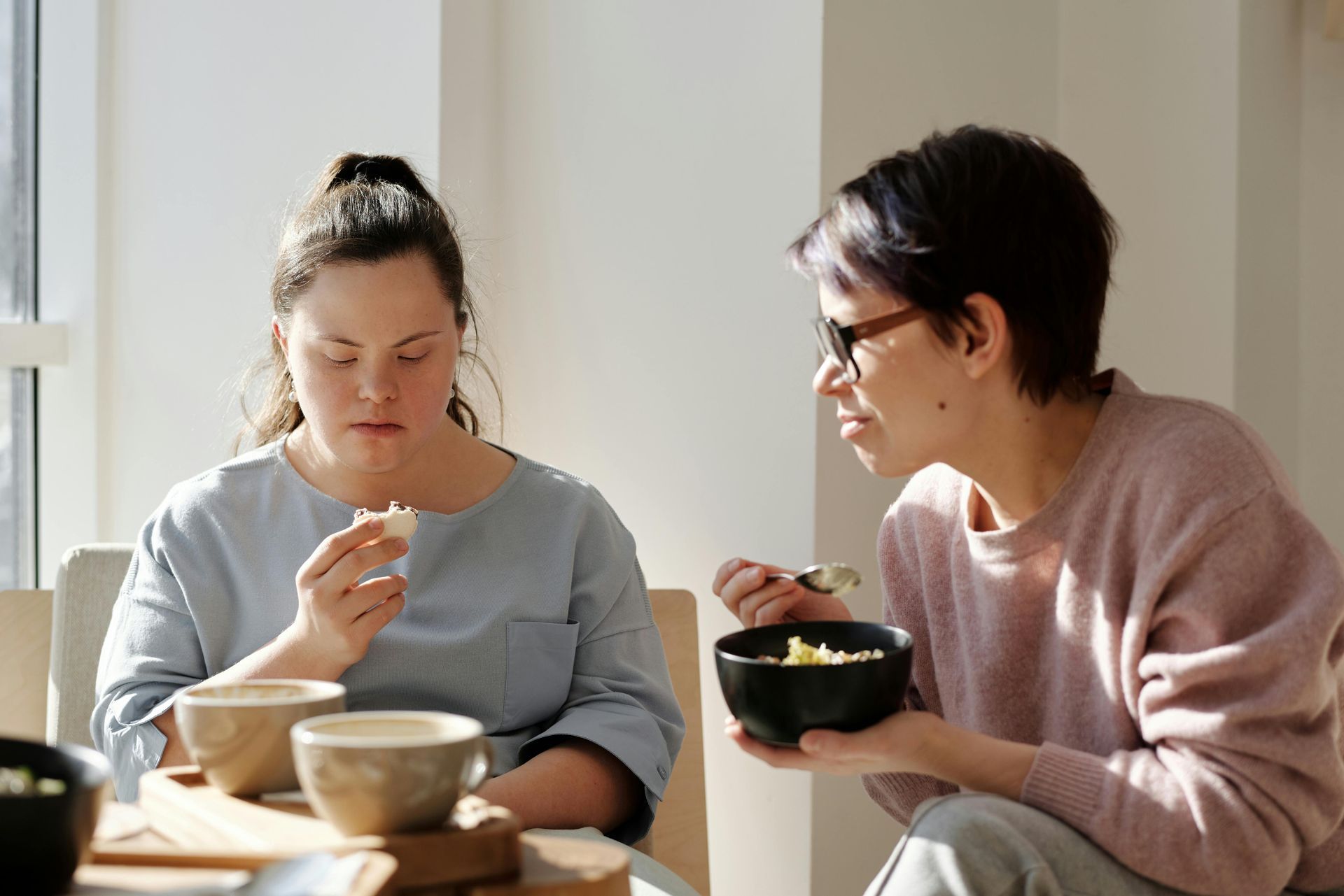 Two people eating at a table, one holding a bowl and spoon in a bright room.