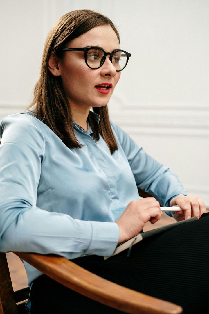 Woman in glasses seated with a notebook, wearing a light blue shirt, speaking in a meeting room
