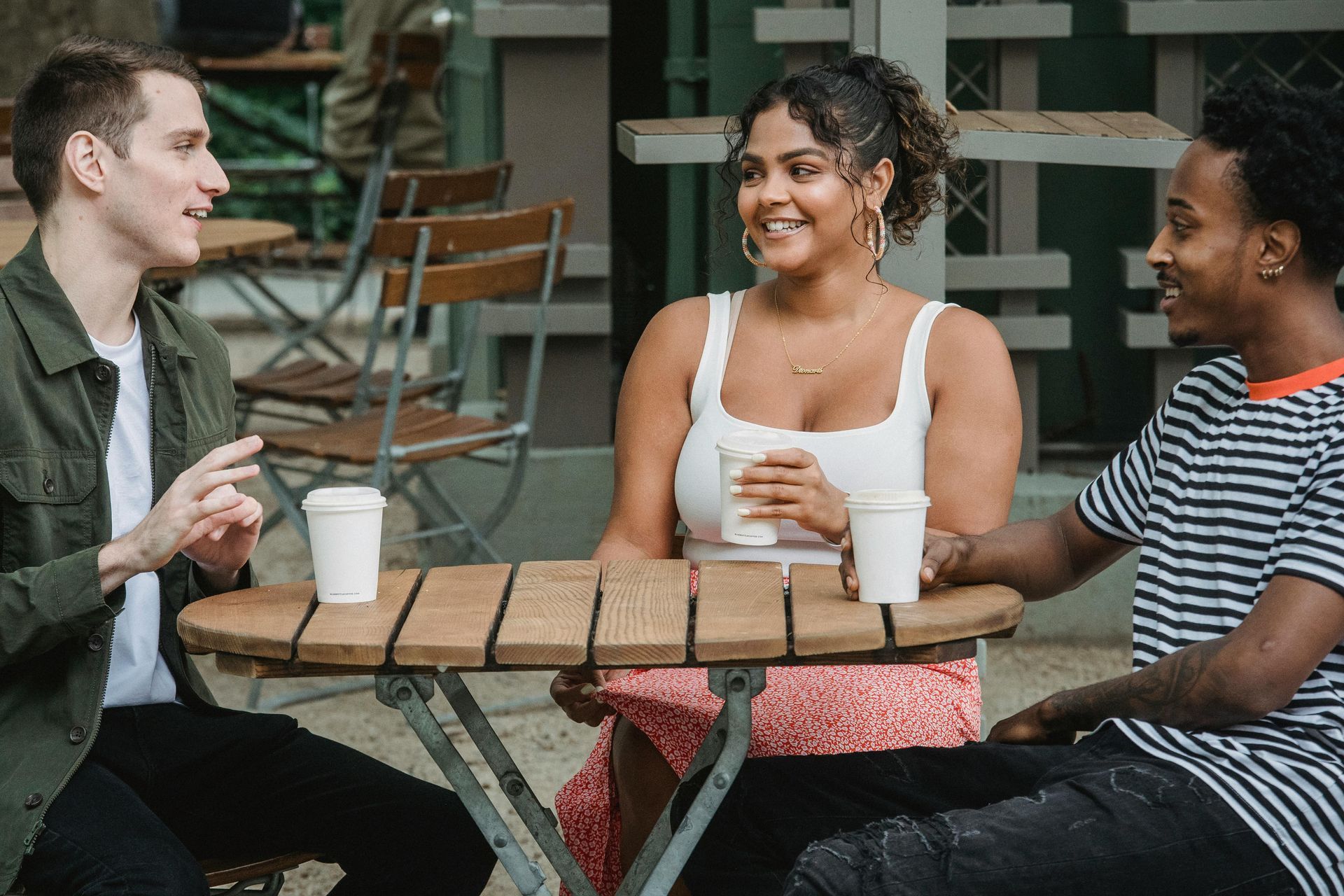 Three people chatting at an outdoor café table with drinks and a laptop