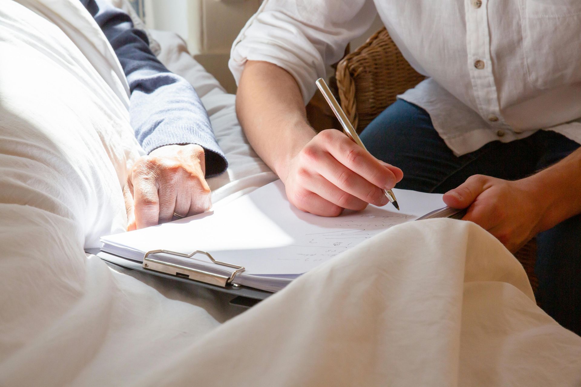 Two people writing on a clipboard under a white blanket in a bright room