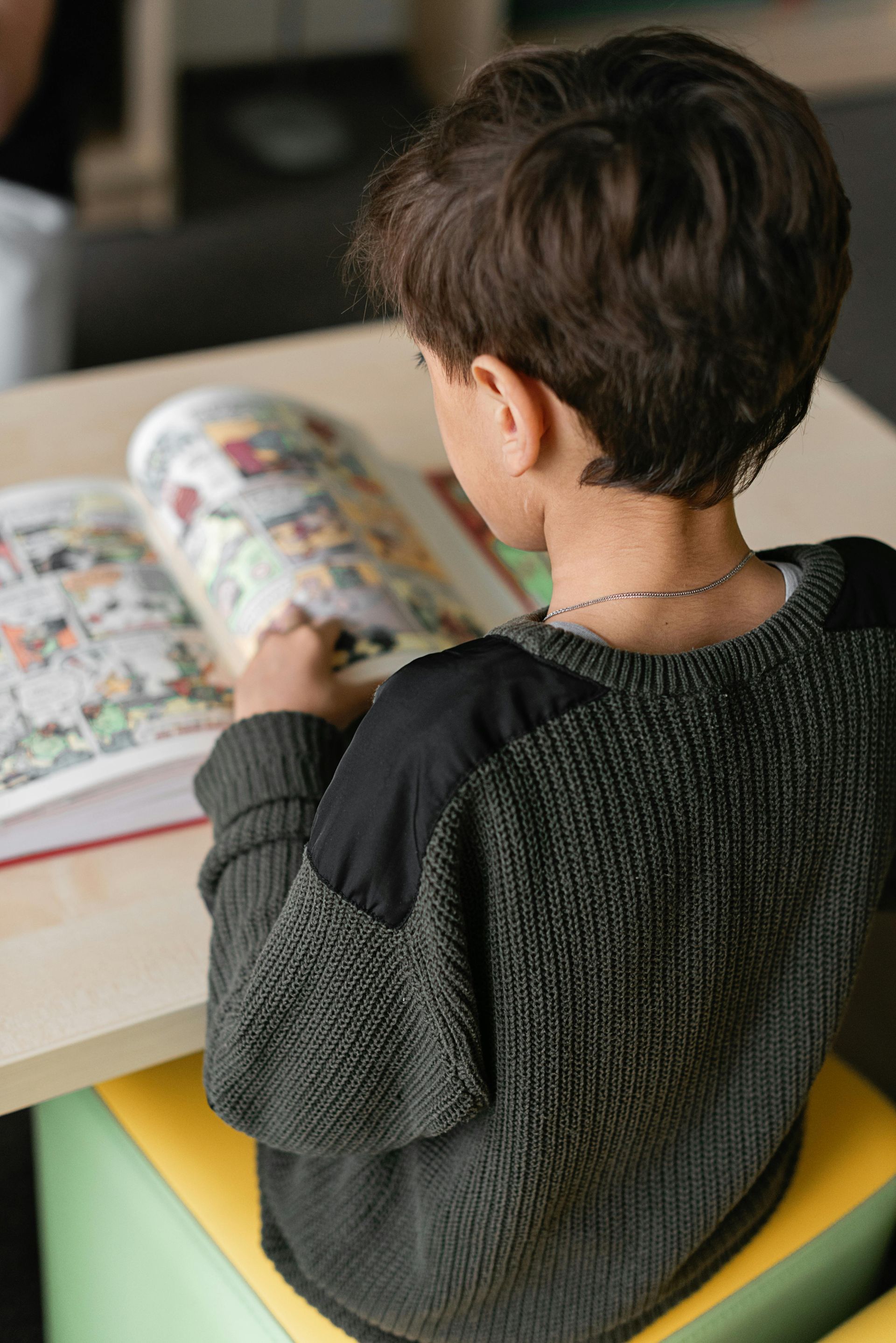 Child reading a colorful magazine at a table, viewed from behind in a cozy indoor setting