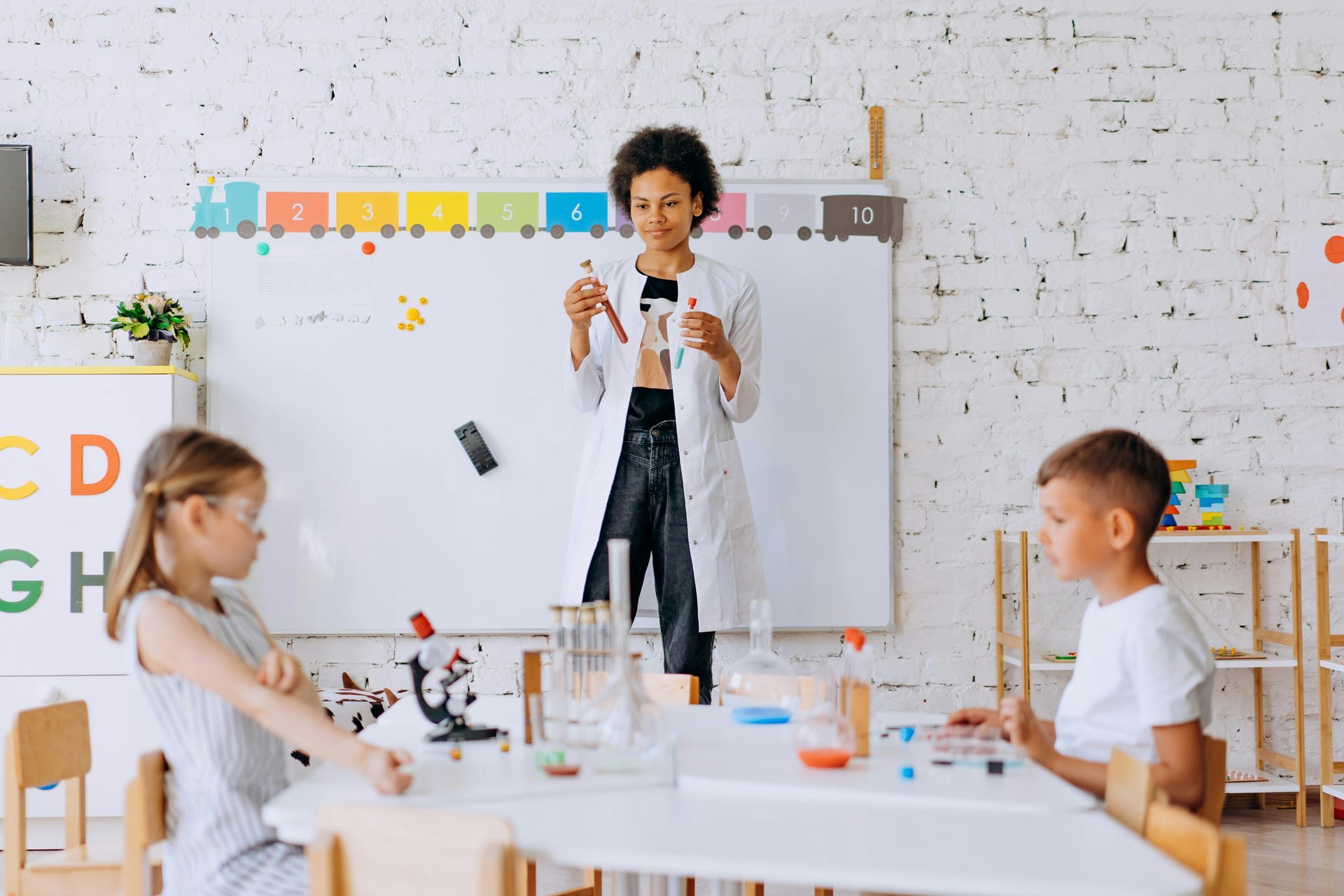 Children doing a science experiment at a classroom table while a teacher holds test tubes near a whiteboard