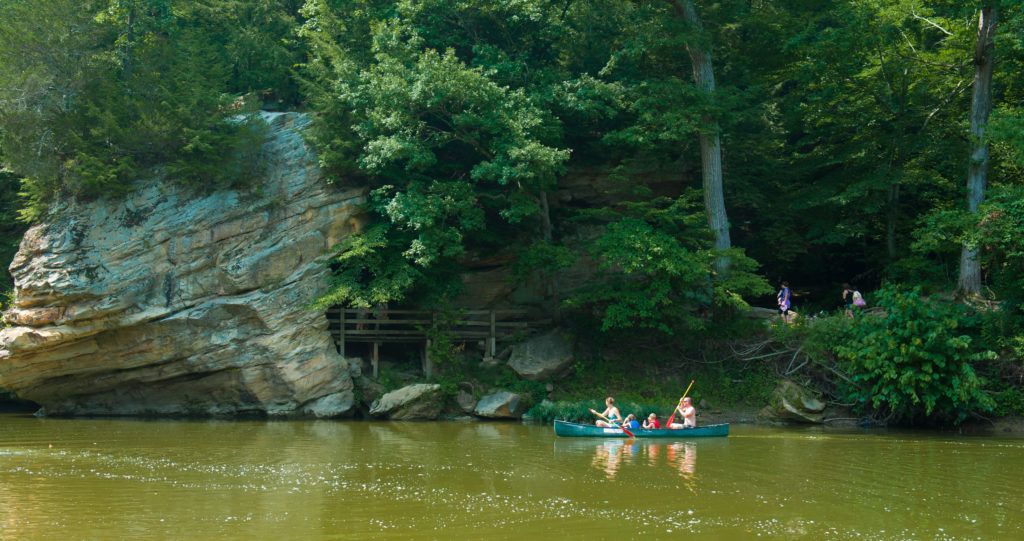Three people paddle a green kayak on a calm river beside a rocky, tree-lined bank.