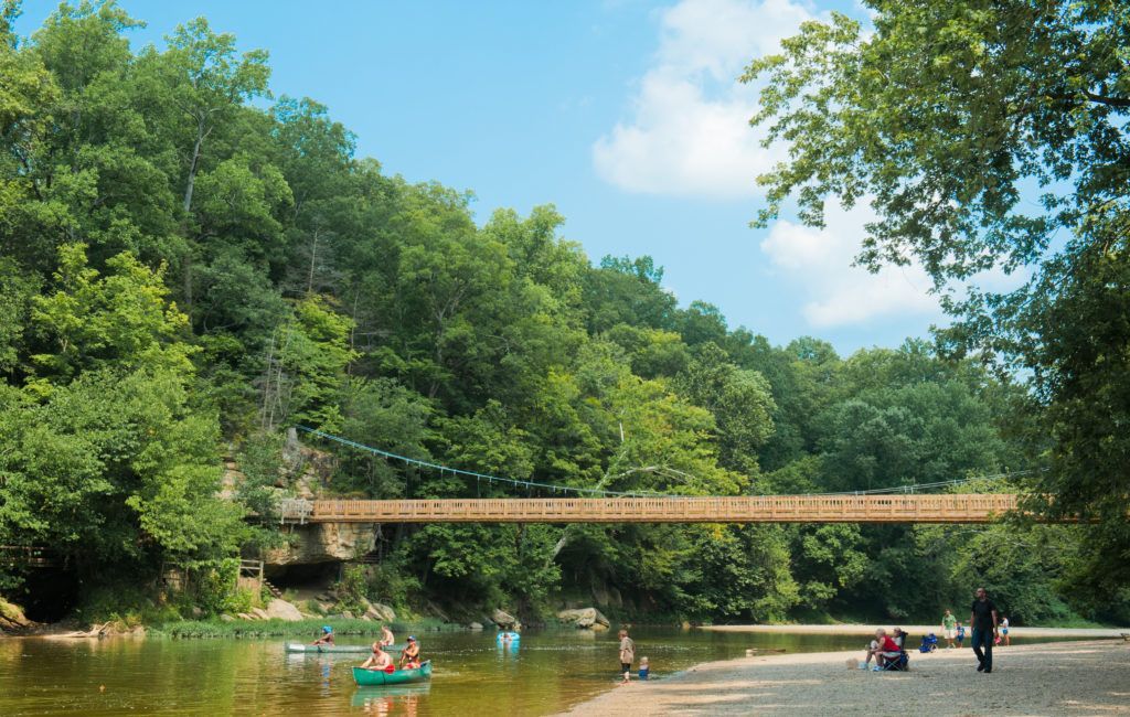 Green riverside scene with a wooden bridge, kayakers, and people walking along a calm shore.