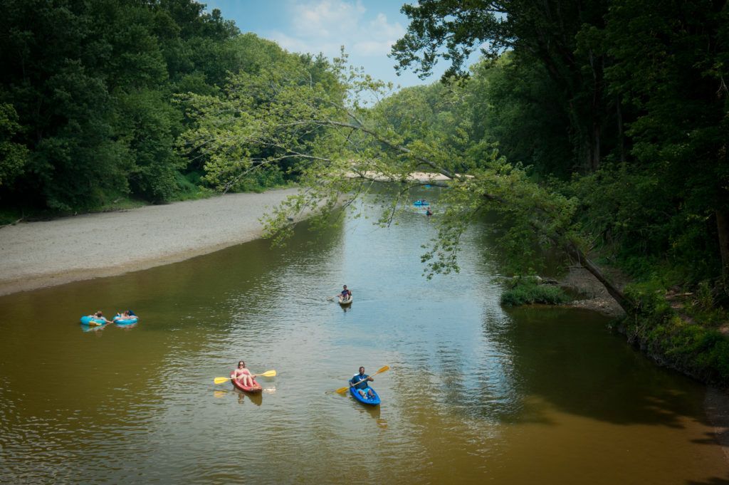 People paddling small blue and red boats on a calm river surrounded by dense green trees