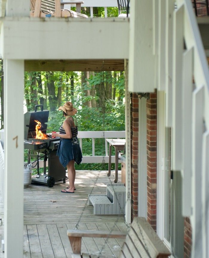 Person grilling on a porch beside a brick house, with white railings and a wooded background.