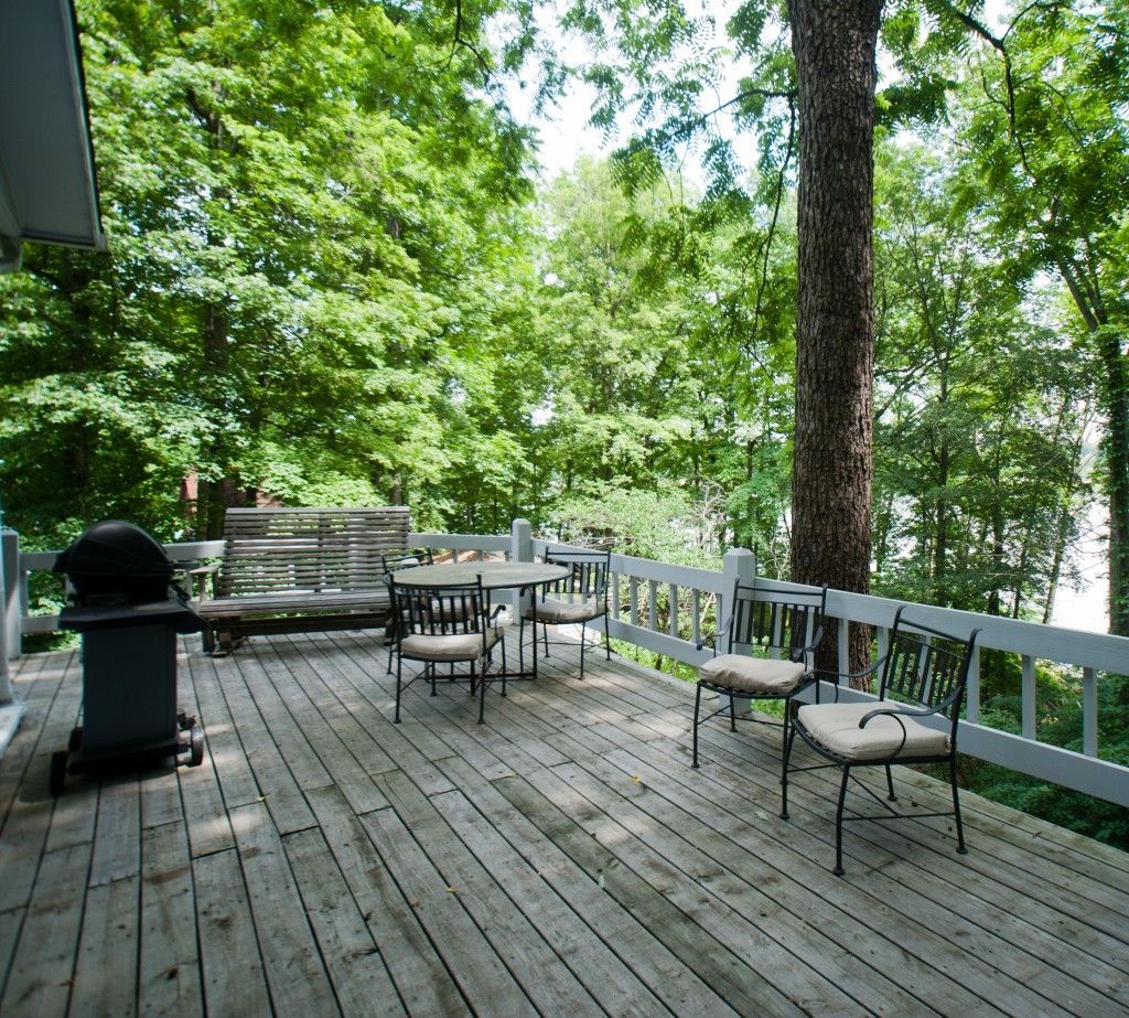 Wooden deck with outdoor chairs and a grill surrounded by lush green trees.