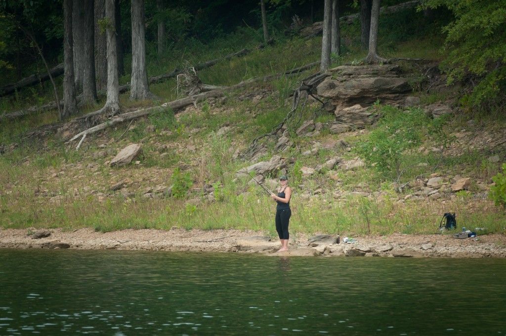Person standing on a rocky lakeshore beside forested trees and green water