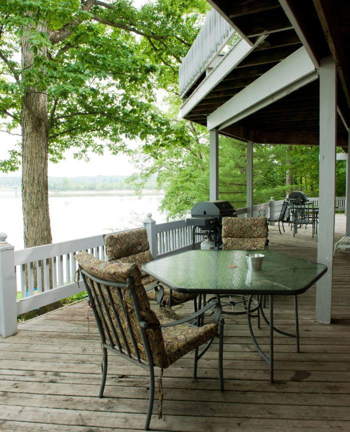 Covered lakeside deck with glass table, chairs, grill, and white railing under green trees.