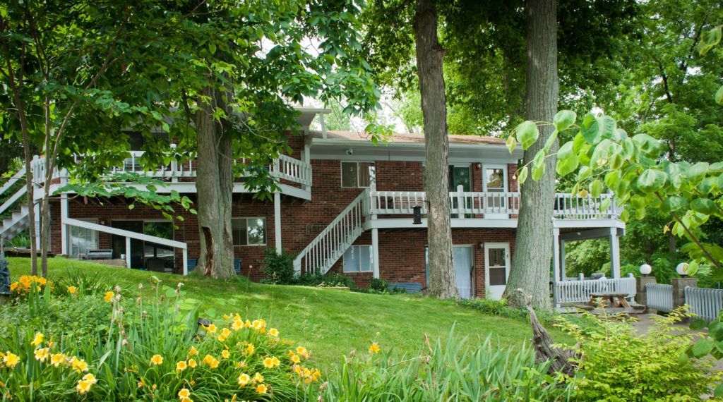 Rustic house with white stairs under tall trees, surrounded by lush green grass and yellow flowers