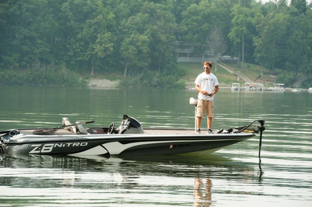 Person standing on a black fishing boat on a calm lake with forested shoreline in the background