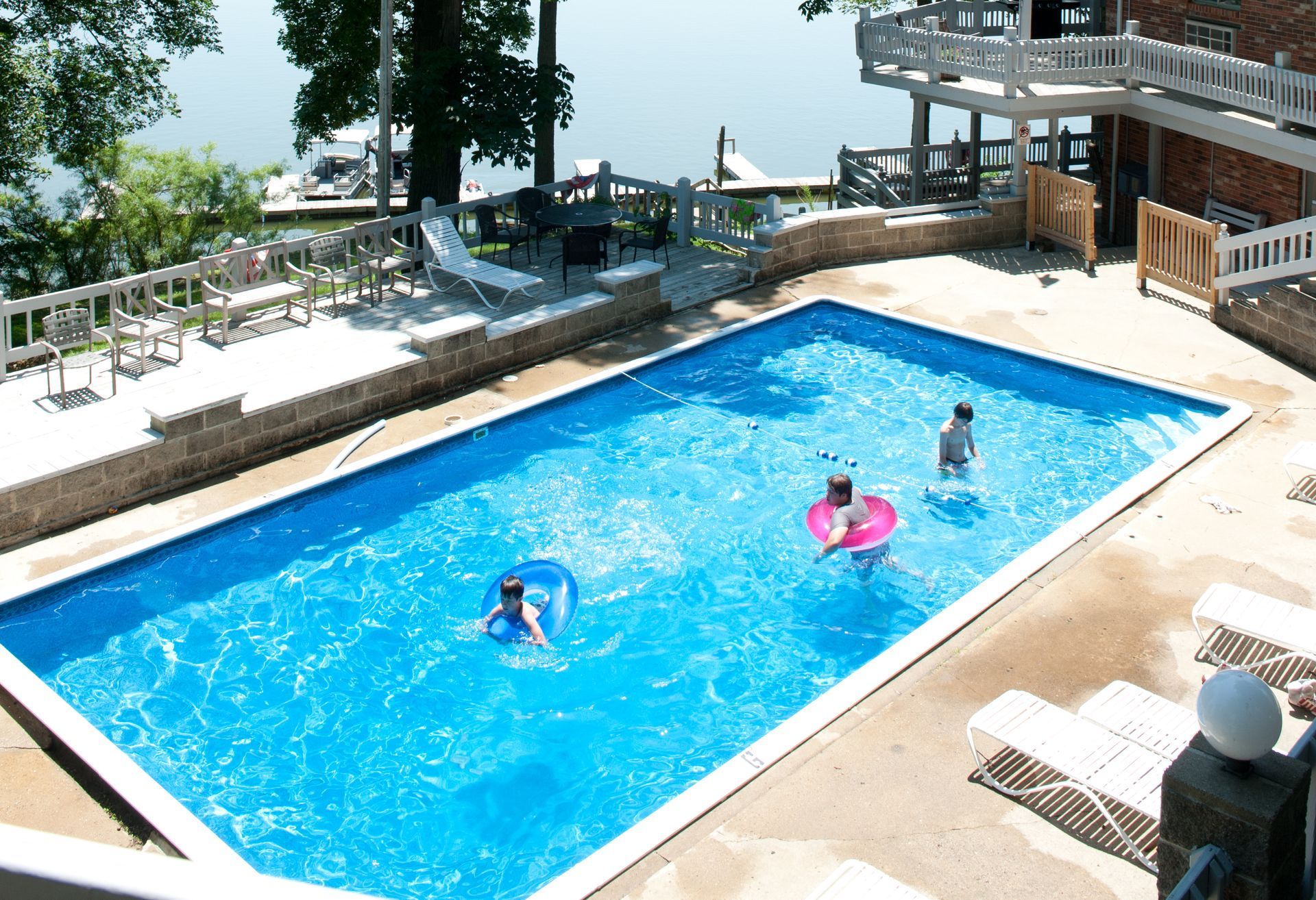 Outdoor swimming pool with three people floating on blue water beside lounge chairs and a lakeside view