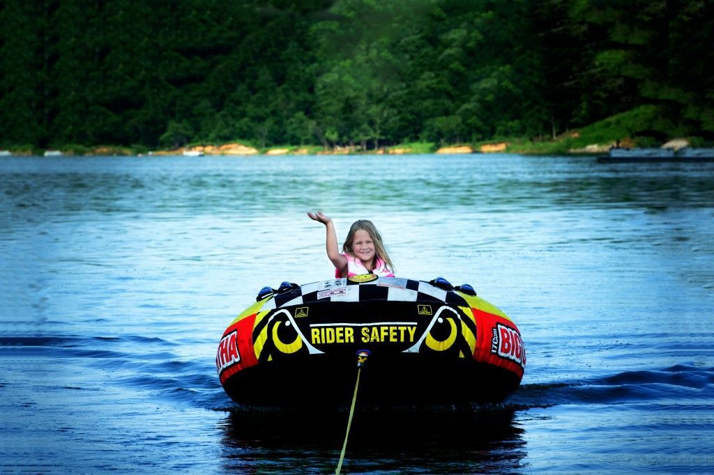 Person waving from a red and black inner tube on a lake, towing behind a boat