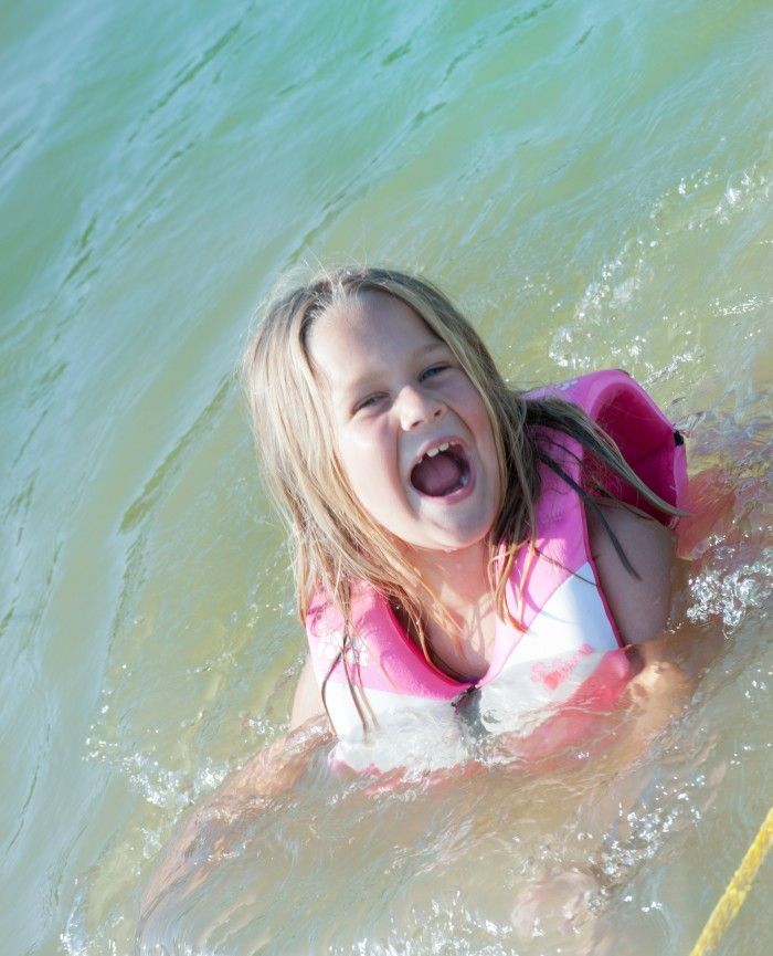 Child in a pink life jacket laughing and splashing in shallow water at the beach.