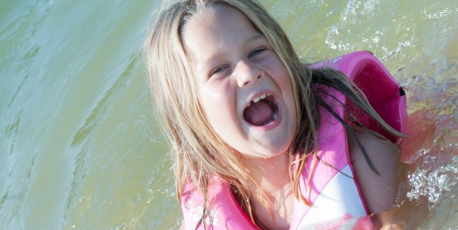Child laughing in green water, wearing a pink life vest and white swimsuit