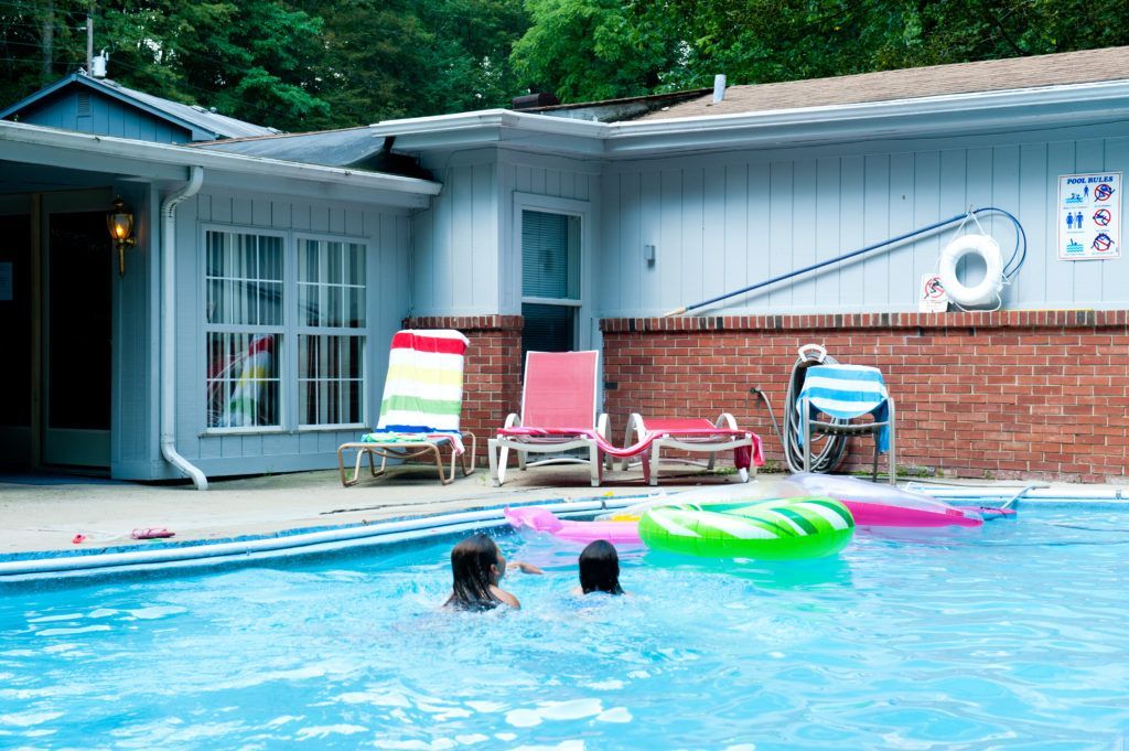 Children swimming in a pool beside lounge chairs and a brick building with a sliding door.