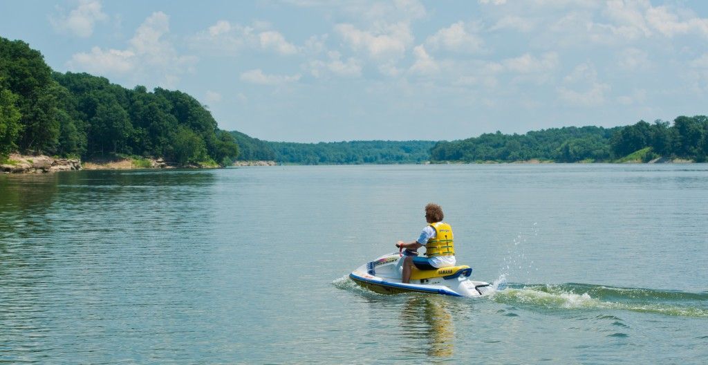 Person riding a jet ski on a calm lake, leaving a small wake near wooded shores.