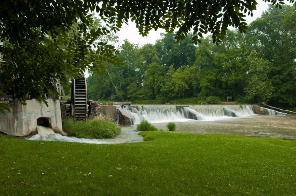 Small waterfall spilling over a low dam into a green park pond with trees overhead