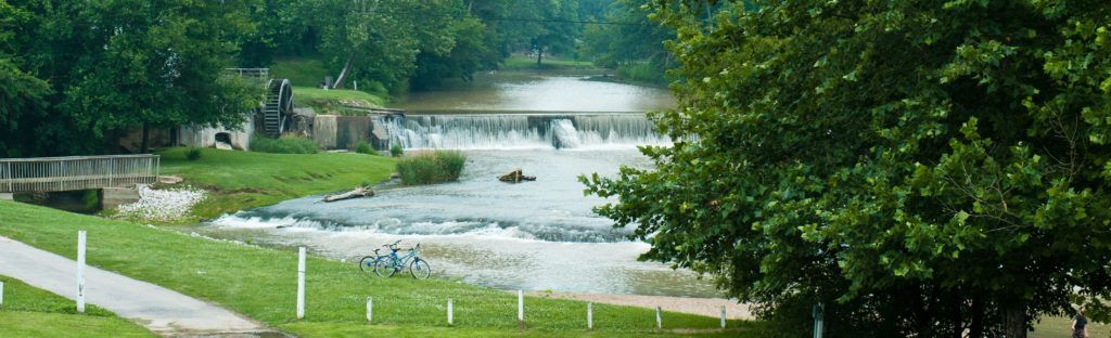 Flooded riverside landscape with a swollen stream, white fence posts, and green trees
