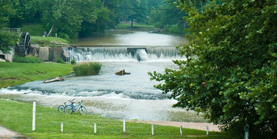 Wooded riverside scene with a low waterfall and fast-flowing water under a cloudy sky