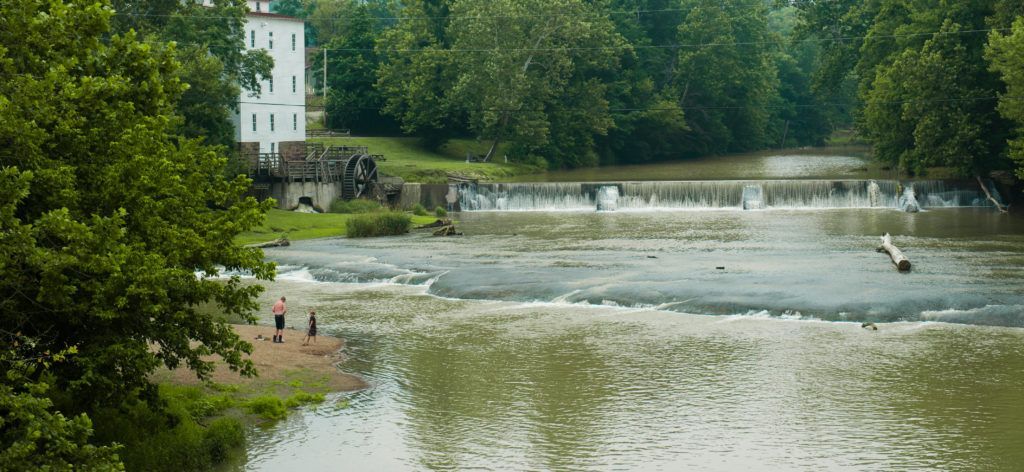 River with a small waterfall beside a white building and green trees