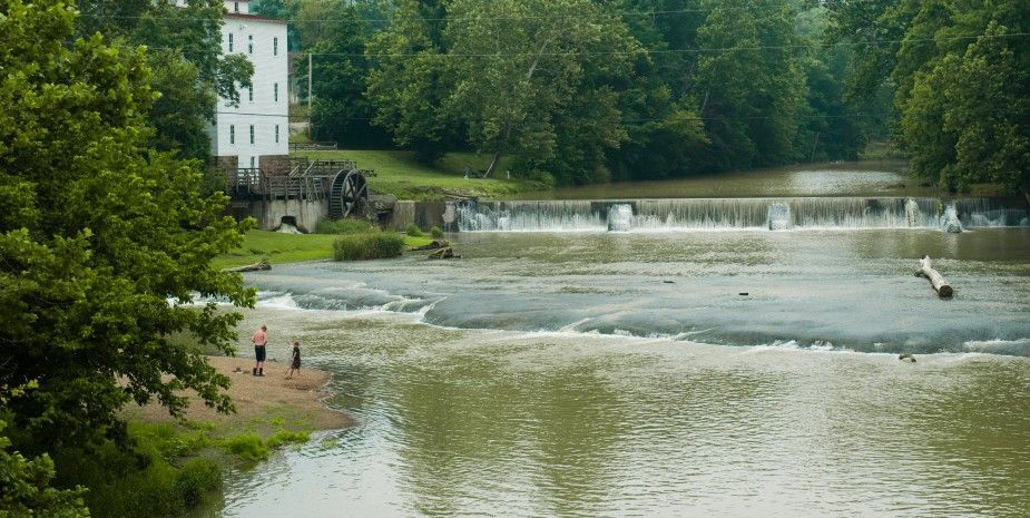 River beside a wooded dam and a white building, with a small waterfall and people on the bank