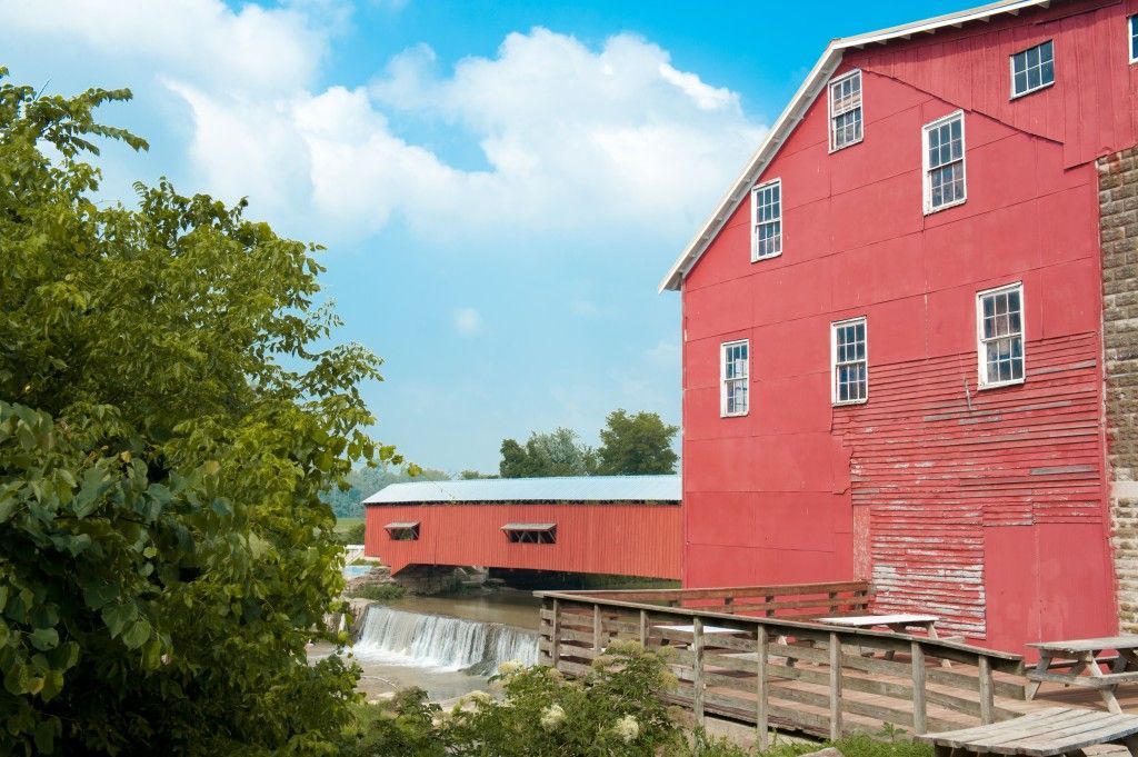 Red mill building beside a river, with a wooden walkway and a blue sky overhead.