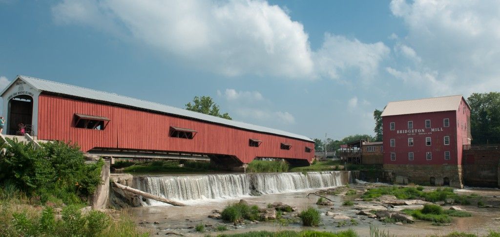 Red covered bridge over a rocky stream beside a red barn under a cloudy sky