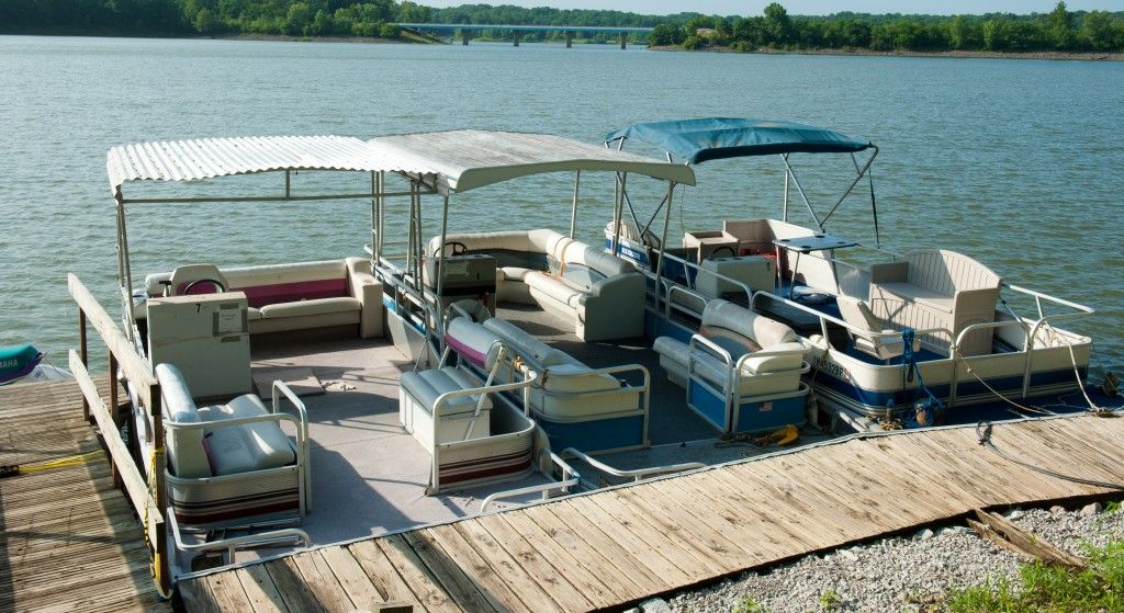 Covered pontoon boats docked on a wooden pier beside a calm lake.