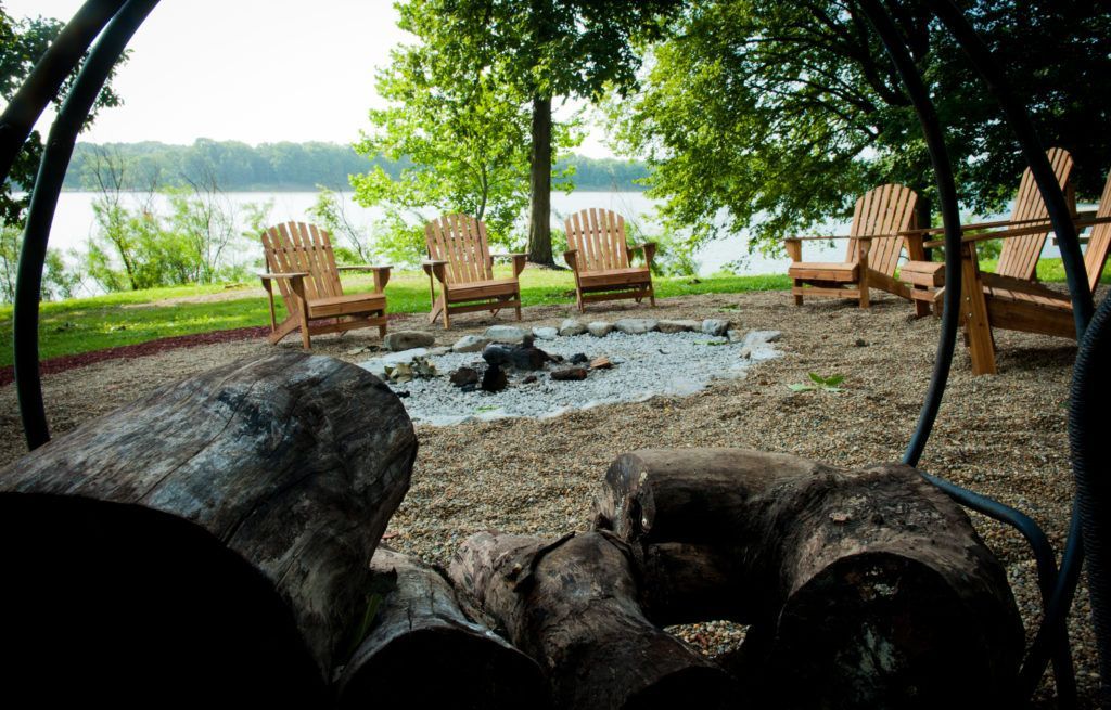 Wooden chairs around a fire pit on a lakeside patio, with rocks and trees in the foreground