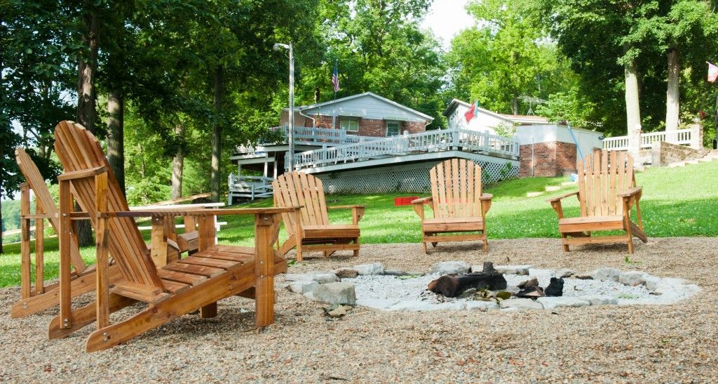 Wooden Adirondack chairs around a stone fire pit on a gravel patio with trees and a house in the background
