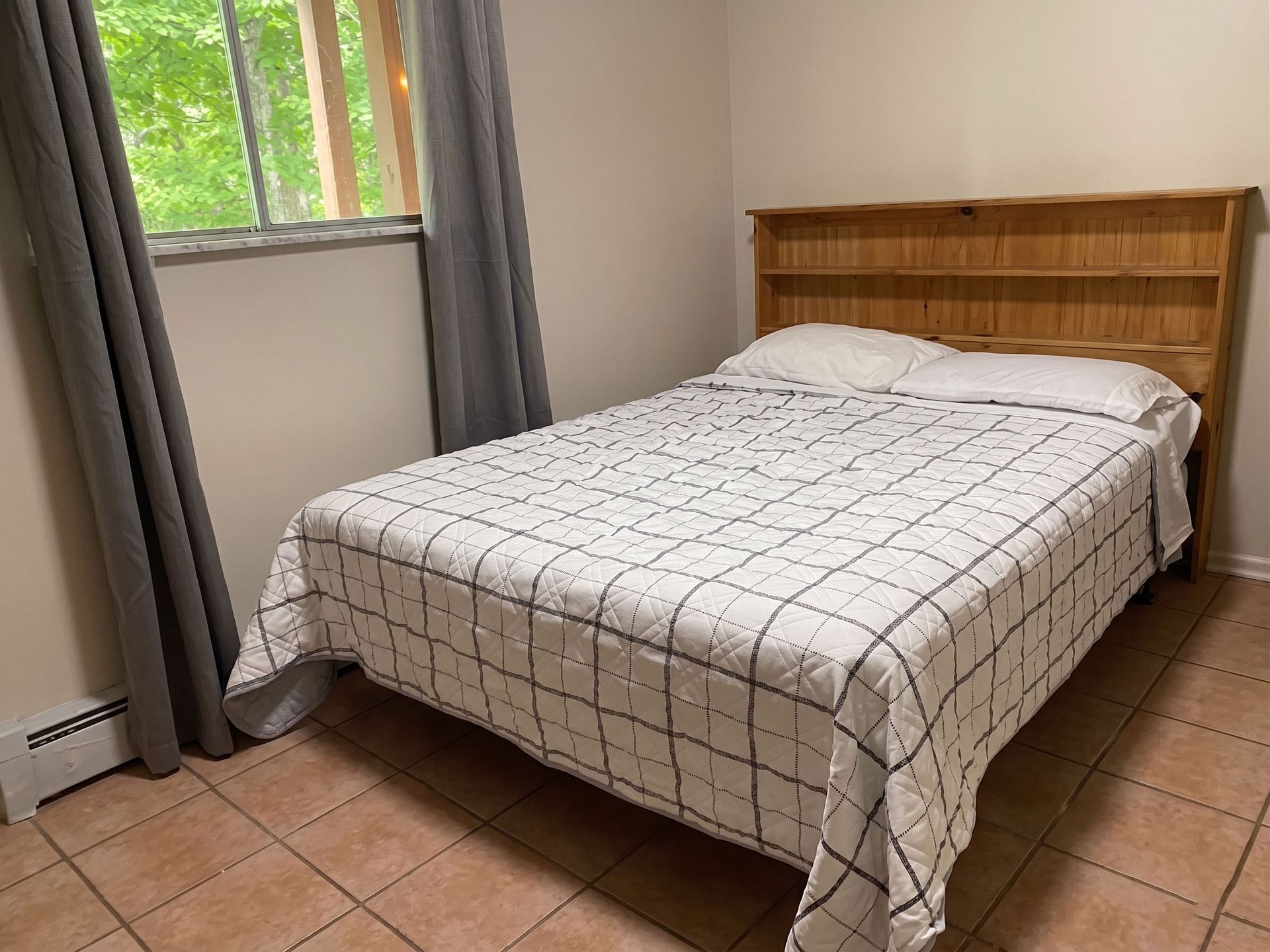 Small bedroom with a bed, wooden headboard, tiled floor, and window with gray curtains.