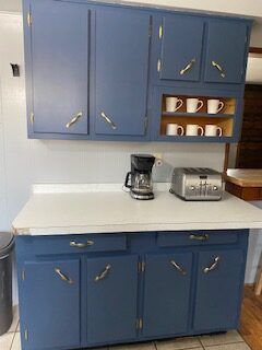 Blue kitchen cabinets with white countertop, coffee maker, toaster, and mugs on a wall shelf.