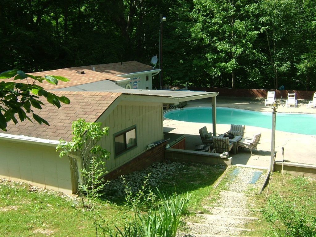 Small house beside a turquoise pool, with patio chairs and trees in a sunny yard