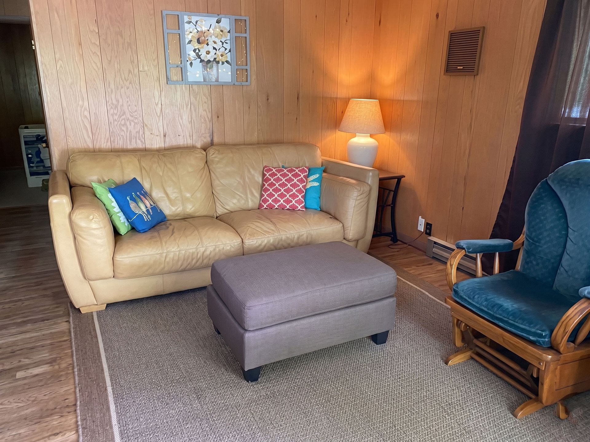 Living room with beige sofa, gray ottoman, blue chair, and lamp against wood-paneled walls.