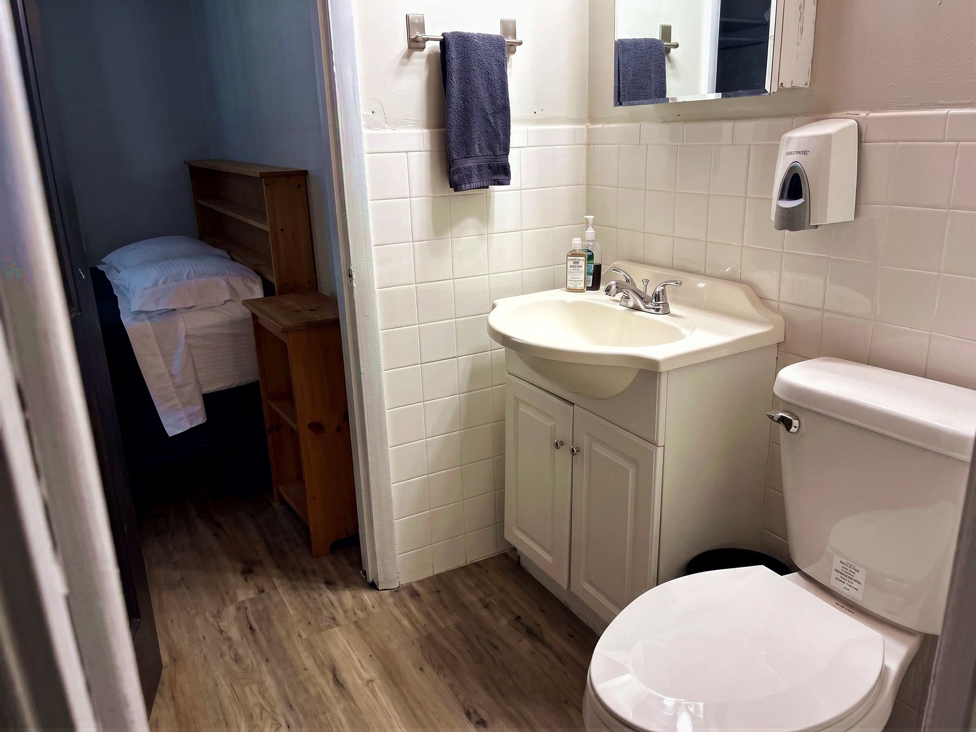 Small bathroom with toilet, sink, towel, and an open doorway to a bedroom with a bed.