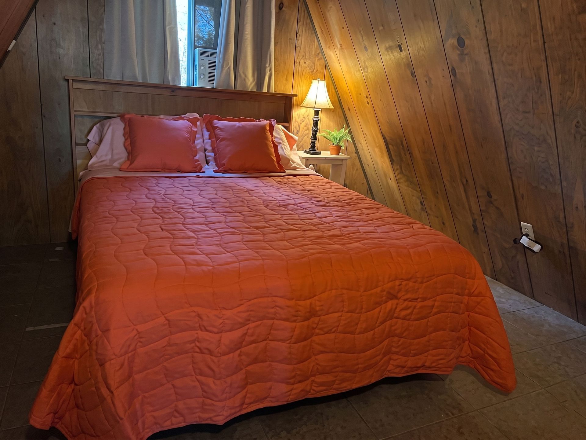 Orange bedspread on a bed in a rustic wood-paneled bedroom with a lit bedside lamp.