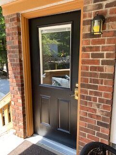 Black front door with glass panel in brick entryway, beside a wall lantern and wooden railing
