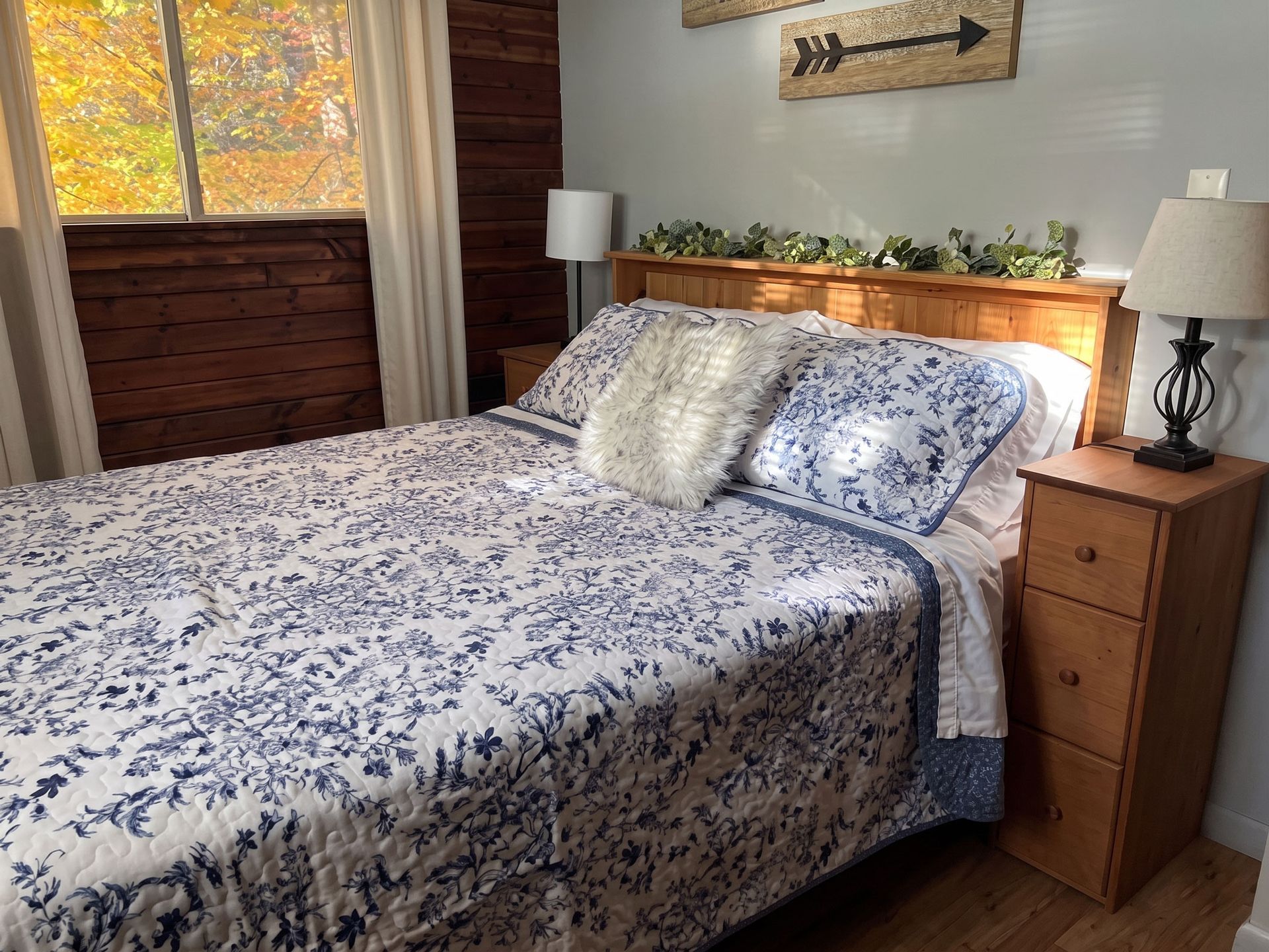 Cozy bedroom with a floral bedspread, wooden nightstands, lamp, and sunlight through the window.