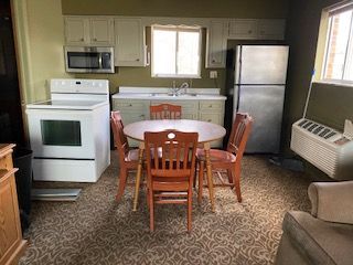 Compact kitchen and dining area with white appliances, round table, wooden chairs, and brown carpet.