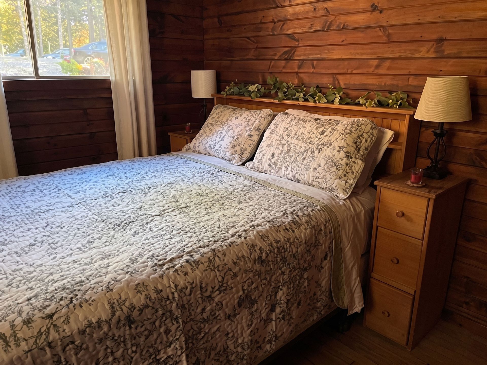 Cozy bedroom with a floral bedspread, wooden walls, bedside tables, lamps, and a window view