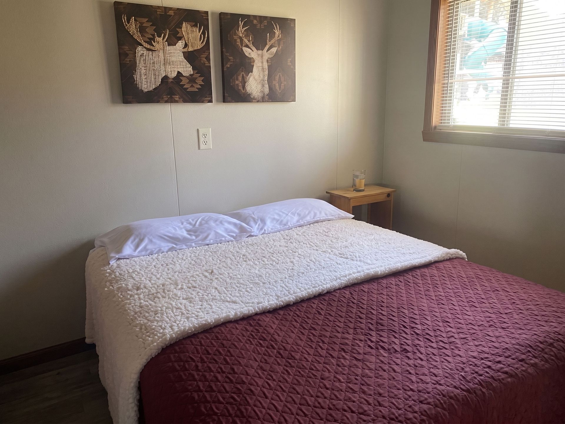 Small bedroom with a made bed, maroon blanket, wall art, and a window with blinds.