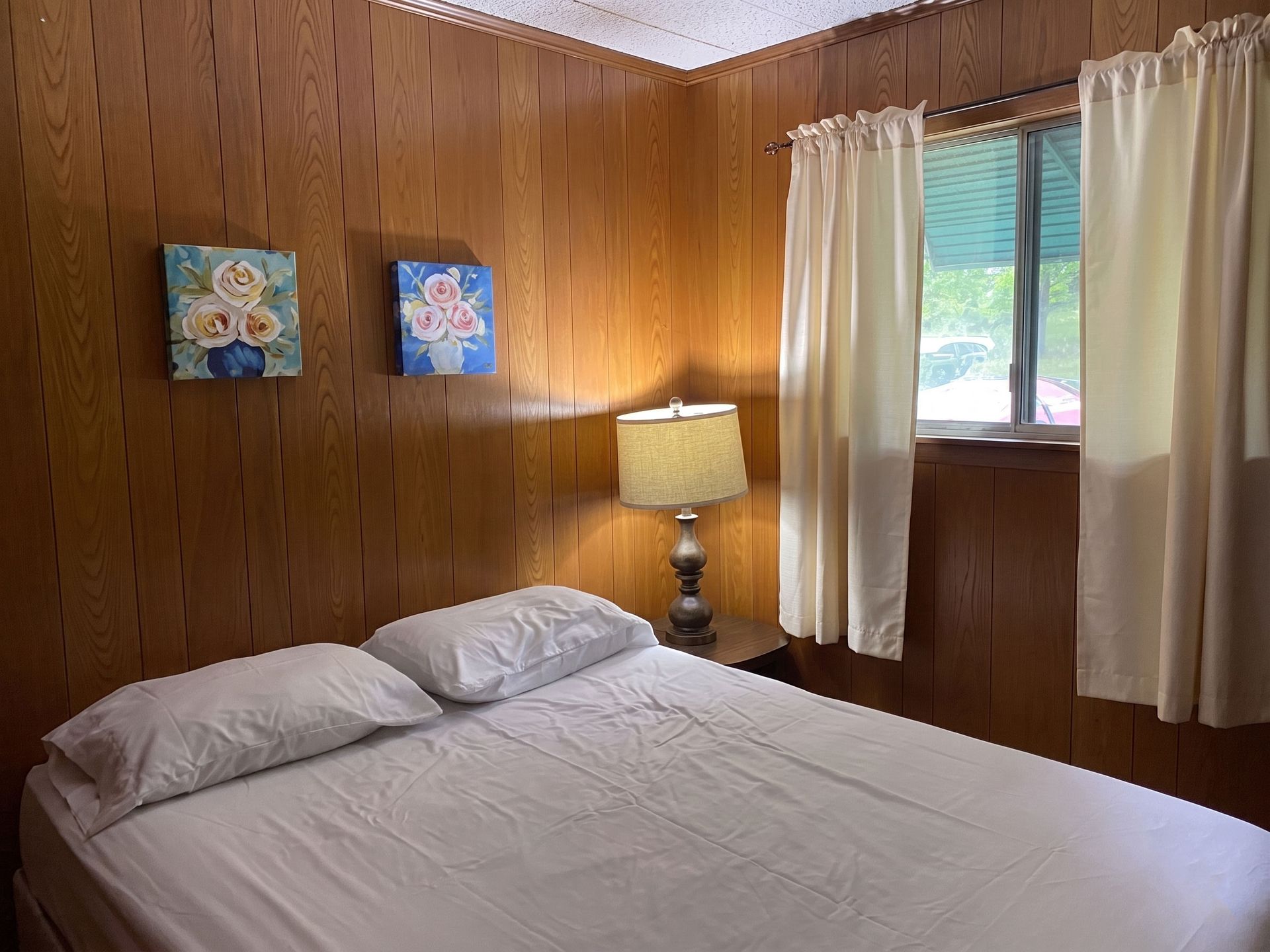 Small wood-paneled bedroom with bed, lamp, wall art, and curtained window