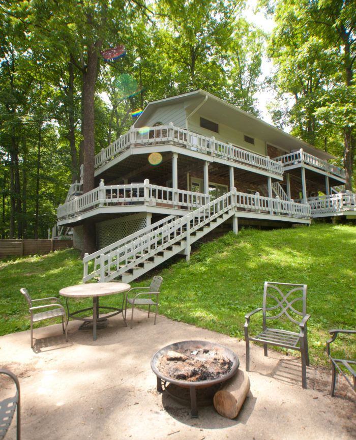 White porch house in a wooded yard with stairs, seating, and a fire pit