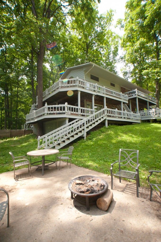 White house with wraparound porch and stairs on a wooded hillside, with a fire pit and chairs in front.